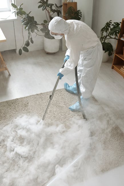 A yellow canister vacuum cleaner with a black hose and a metal wand attachment is positioned on a patterned area rug in a room with wooden flooring. To the left, a black storage unit with a wooden top